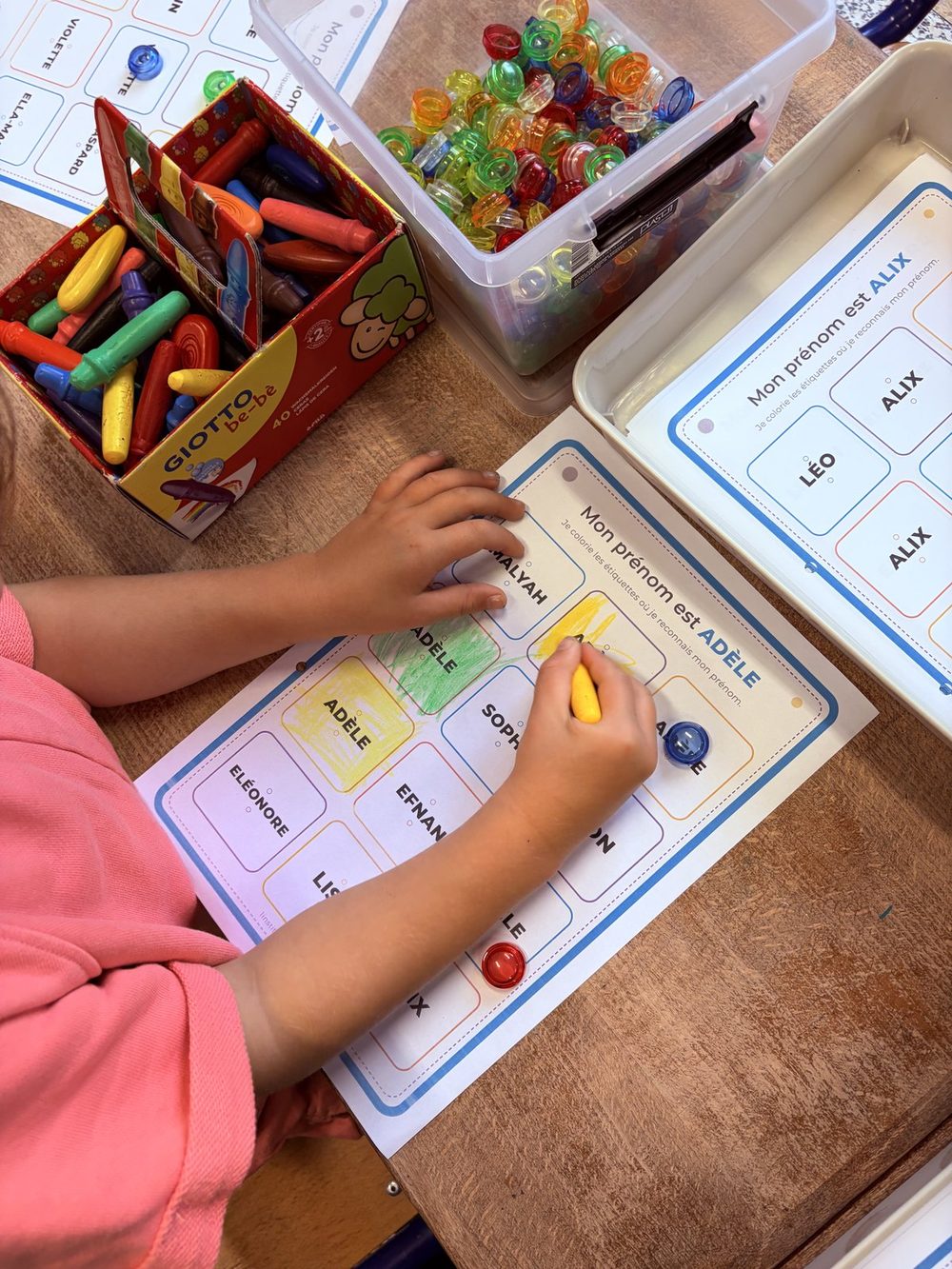 Un enfant colorie aux crayons de couleur les étiquettes portant son prénom